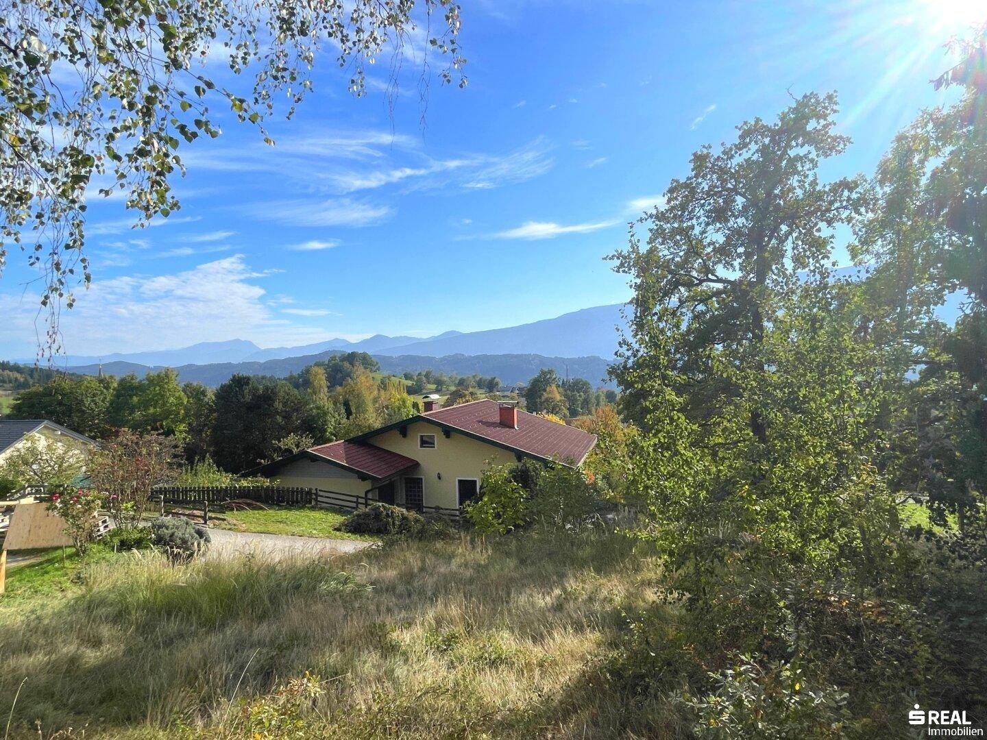 Haus mit rotem Dach in sonniger Lage, umgeben von Bäumen und mit weitem Blick auf die Berglandschaft.