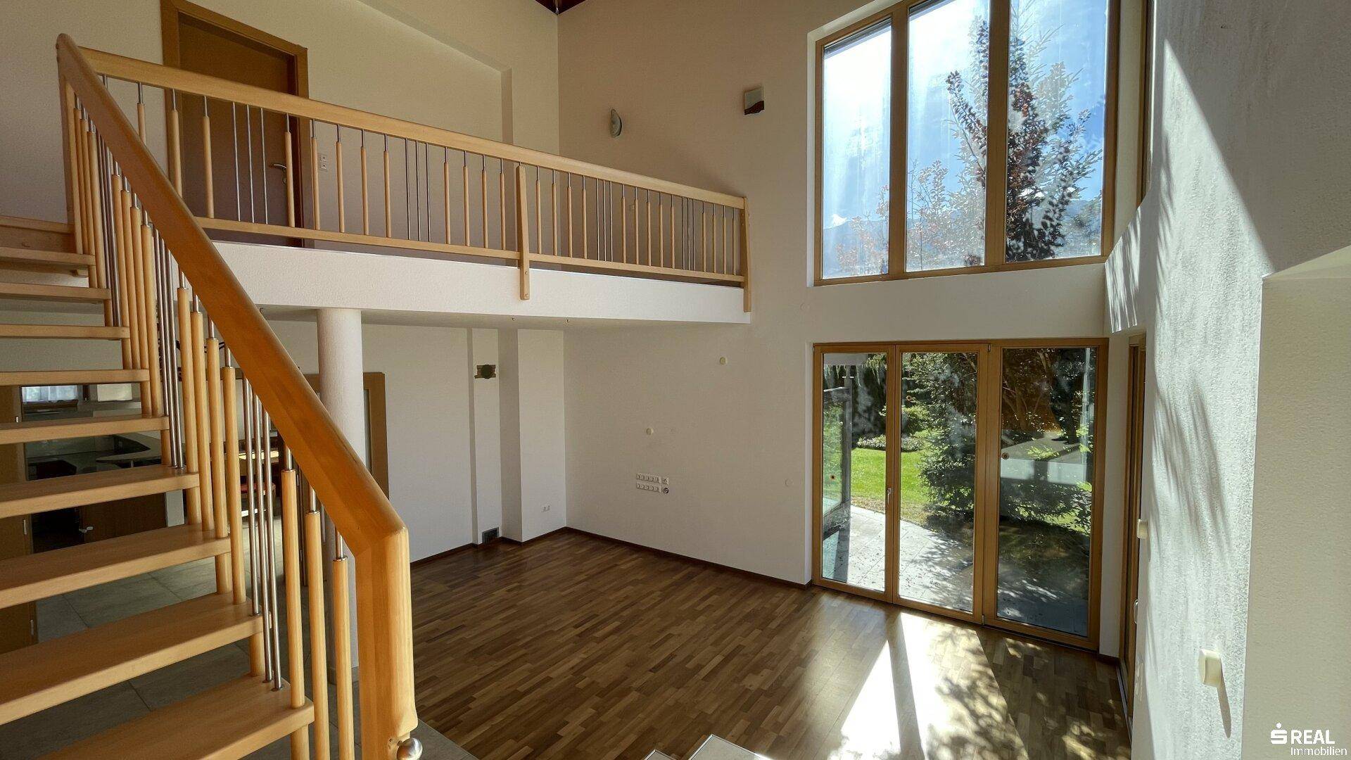 Geräumiger Wohnbereich mit Holztreppe, Galerie und großen Fenstern, die viel Licht hereinlassen.