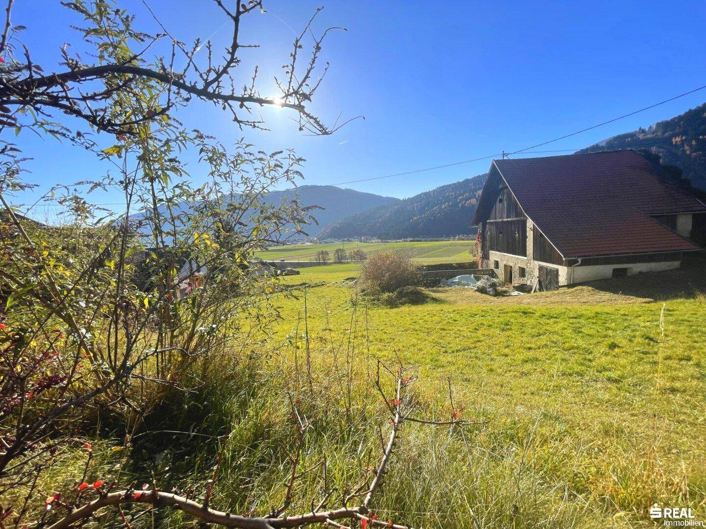 Ländliche Szene mit einem traditionellen Gebäude, weiten Feldern und Bergen im Sonnenlicht.