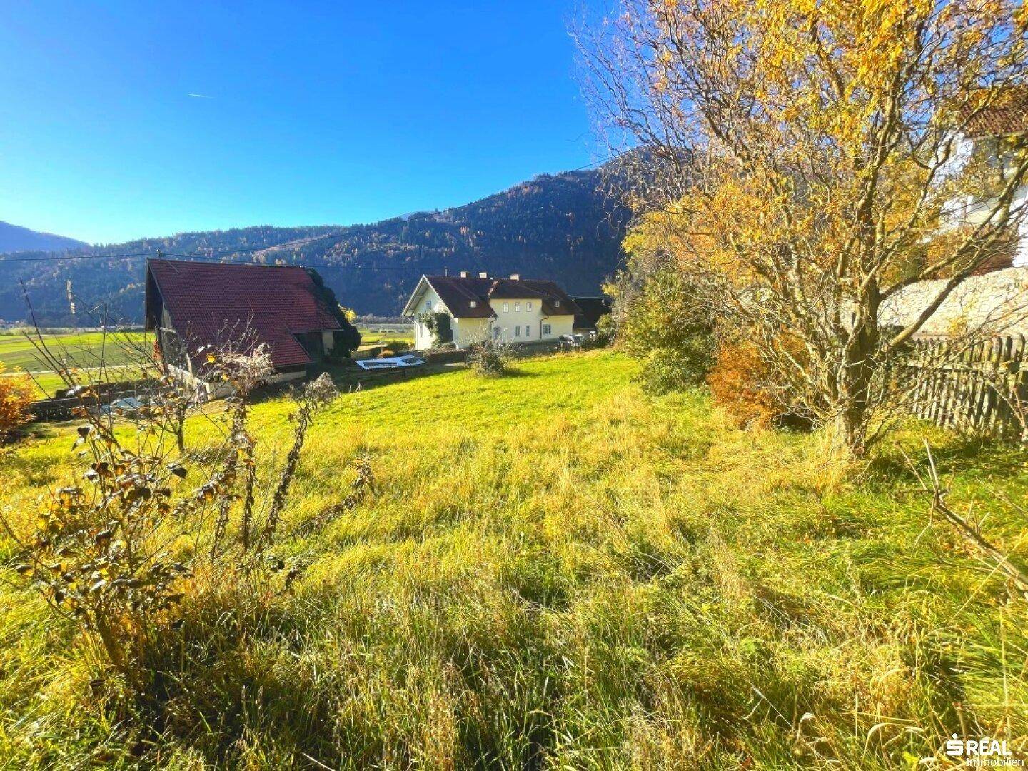 Weitläufige grüne Wiesen mit Häusern im Hintergrund und Bergen unter klarem Himmel.