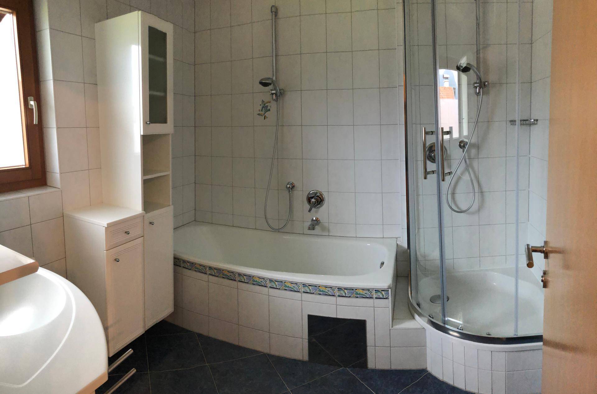 Badezimmer mit weißen Fliesen, einer Eckbadewanne, separater Duschkabine und einem Fenster.