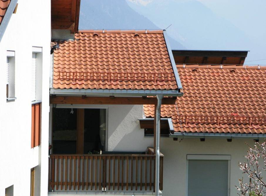 Balkon mit Holzgeländer an einem Haus mit Ziegeldach und Bergblick im Hintergrund.