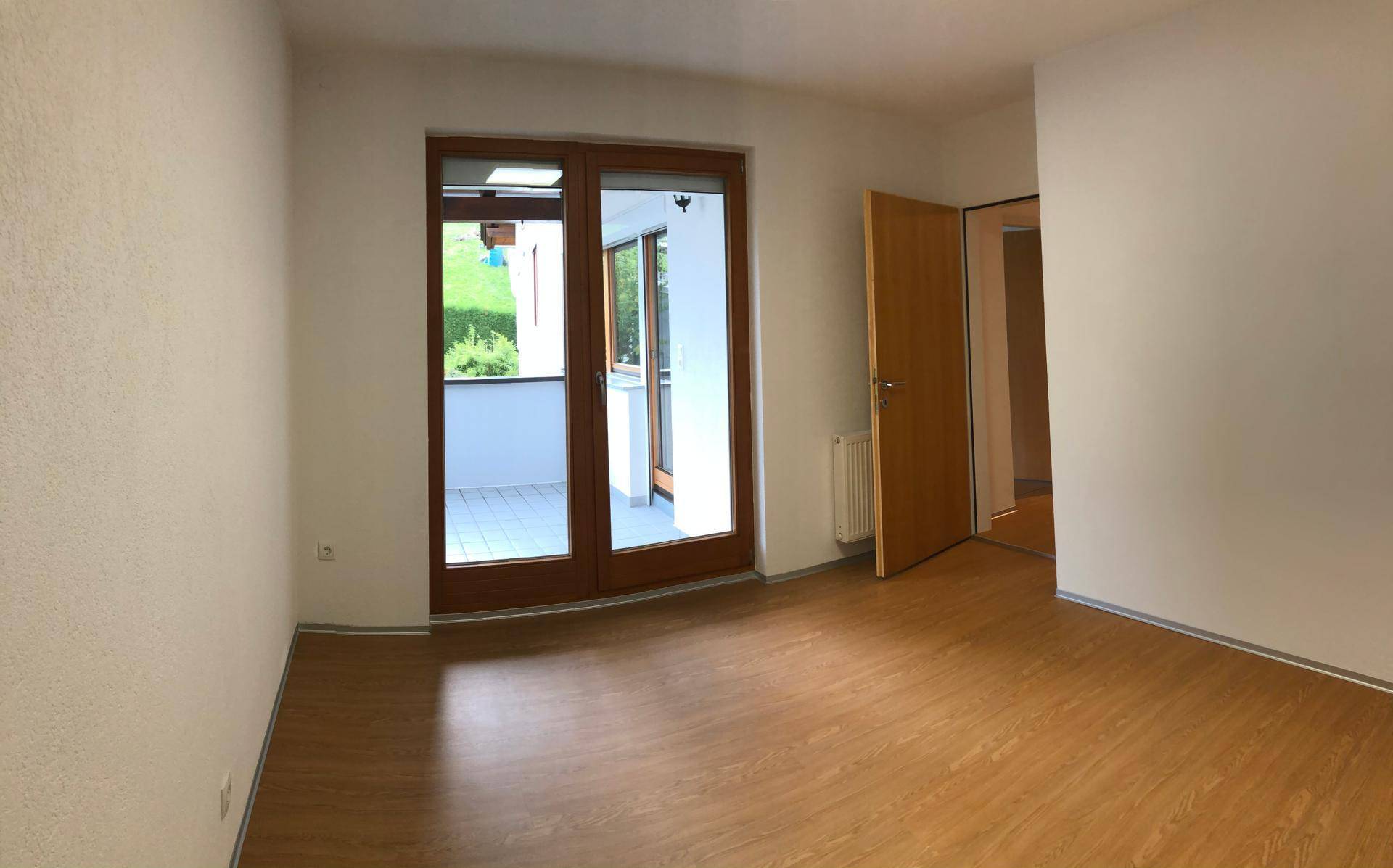 Heller Raum mit Holzboden und großen Glastüren, die zu einem sonnigen Balkon führen.