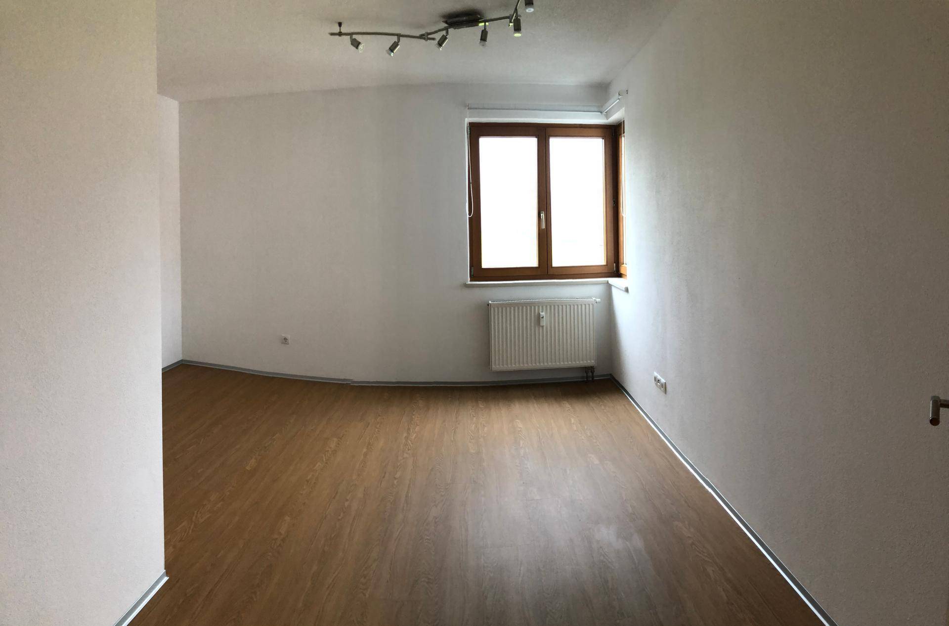 Leeres Zimmer mit hellem Holzboden, weiß gestrichenen Wänden und einem großen Fenster.