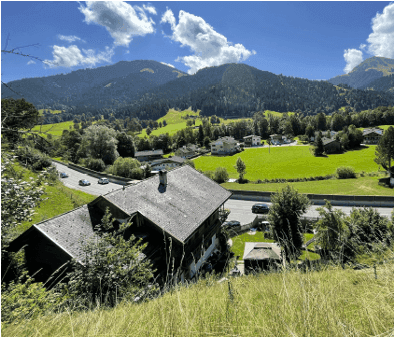 Traditionelles Haus in alpiner Landschaft mit grünen Hügeln und Bergen unter blauem Himmel.