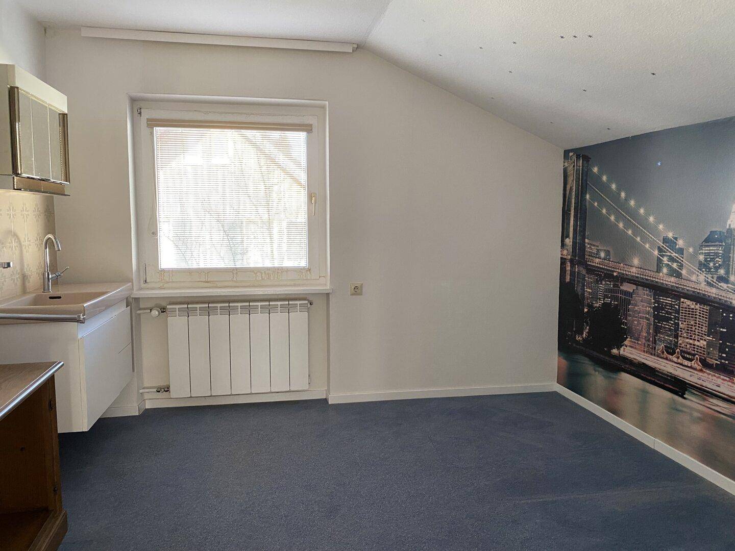 Dachzimmer mit Teppichboden, Fenster und einer kleinen Küchenzeile an der Wand.