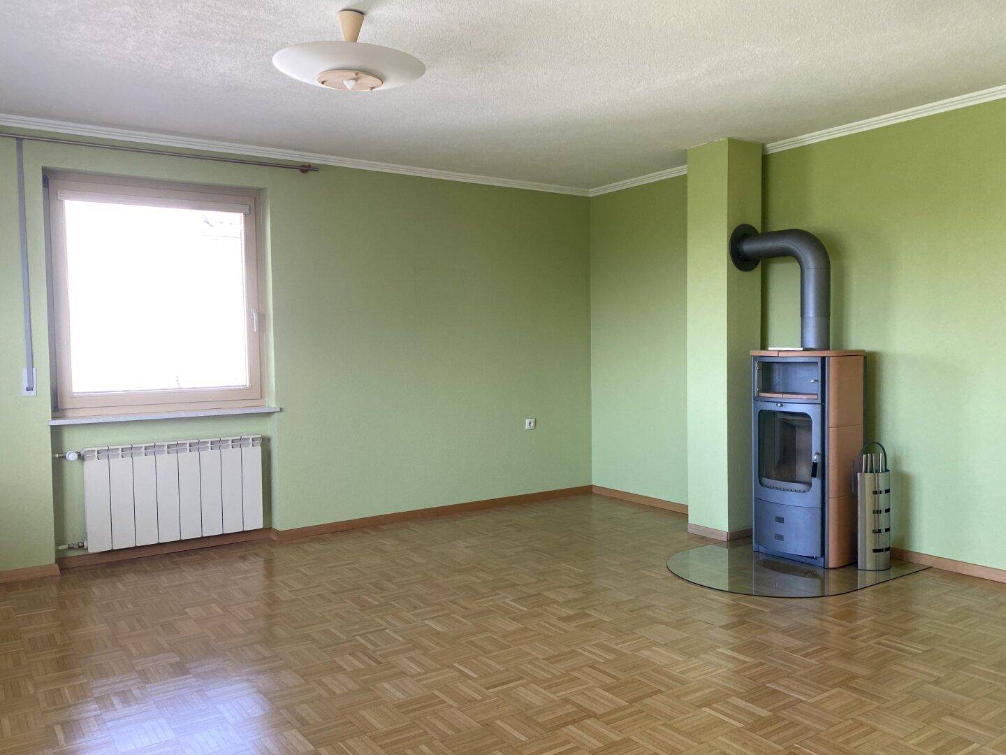Helles Wohnzimmer mit Kaminofen, Parkettboden und Fenster, das viel Tageslicht hereinlässt.
