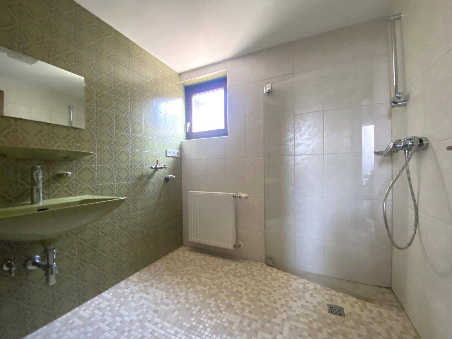 Badezimmer mit Dusche, Waschbecken und Fenster, gefliest in hellgrünen und weißen Tönen.