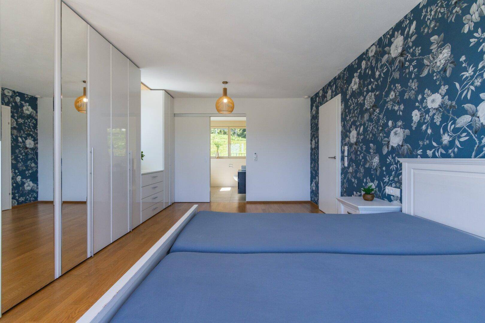 Schlafzimmer mit blauer Blumentapete, Holzboden und Zugang zu einem weiteren Raum.