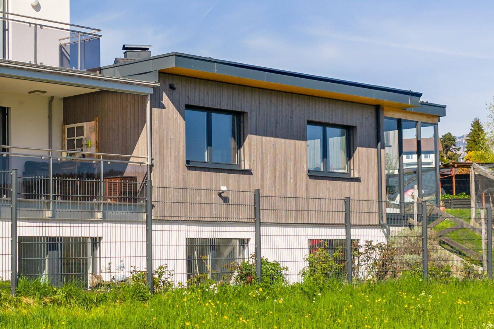 Seitenansicht des Gebäudes mit Holzfassade und einem Balkon mit Glasgeländer.