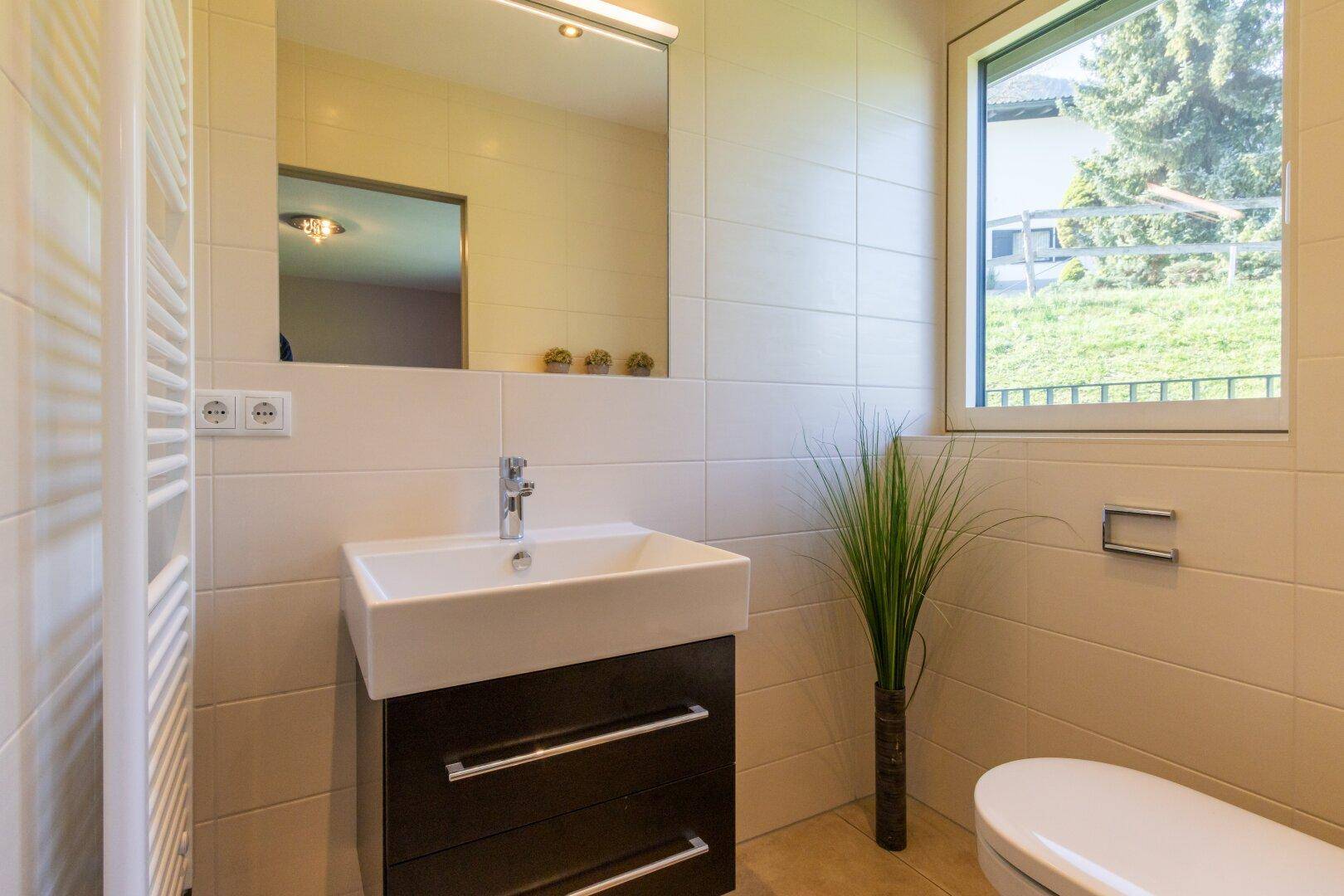 Modernes Badezimmer mit hellen Fliesen, Waschtisch, beheiztem Handtuchhalter und Fenster.
