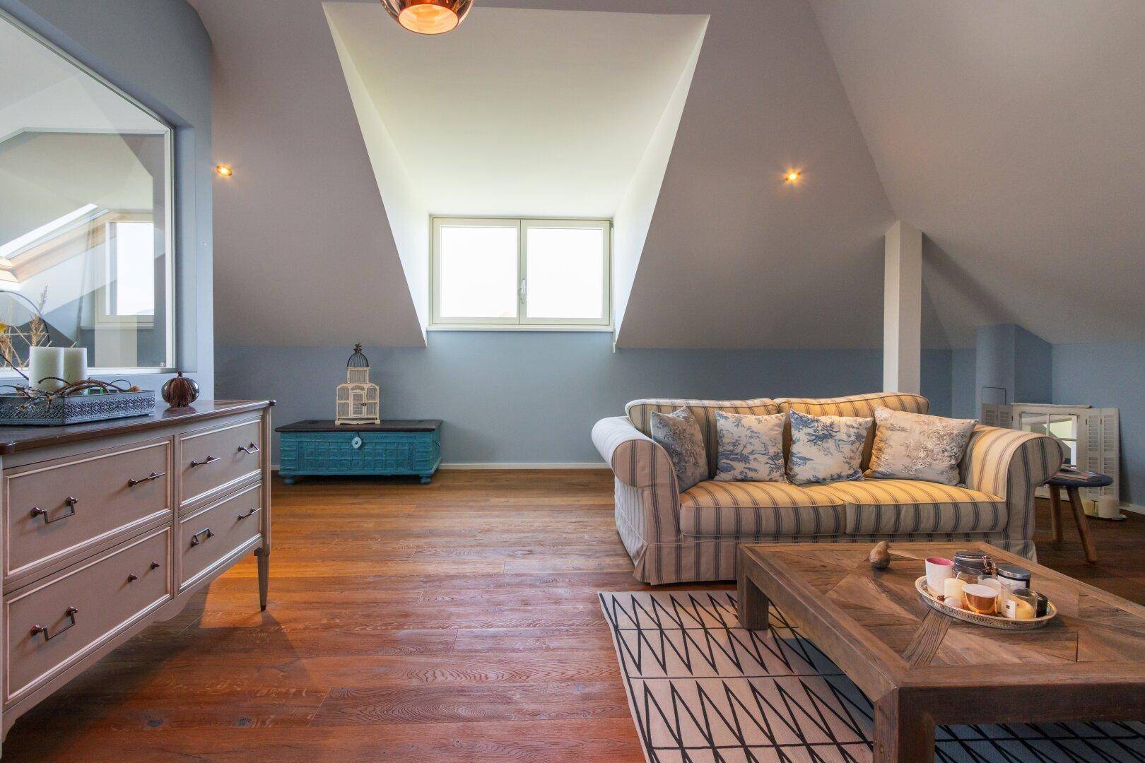 Helles Dachgeschoss-Wohnzimmer mit bequemer Couch, Holzboden und Fenster in der Dachschräge.
