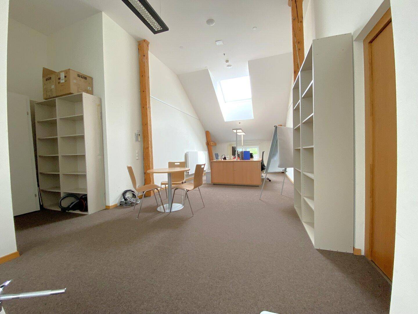 Geräumiges Büro im Dachgeschoss mit Teppichboden, Regalen und einem Schreibtischbereich unter einem Dachfenster.