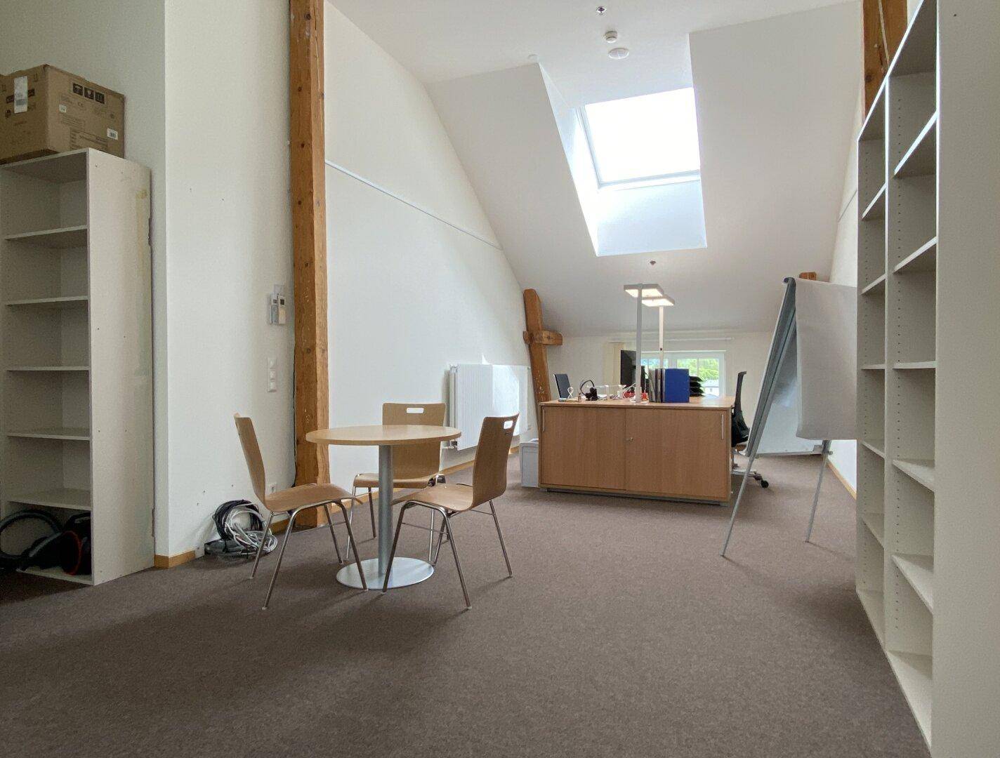 Bürofläche im Dachgeschoss mit Teppichboden, Regalen und einem Arbeitsbereich, beleuchtet durch ein Dachfenster.