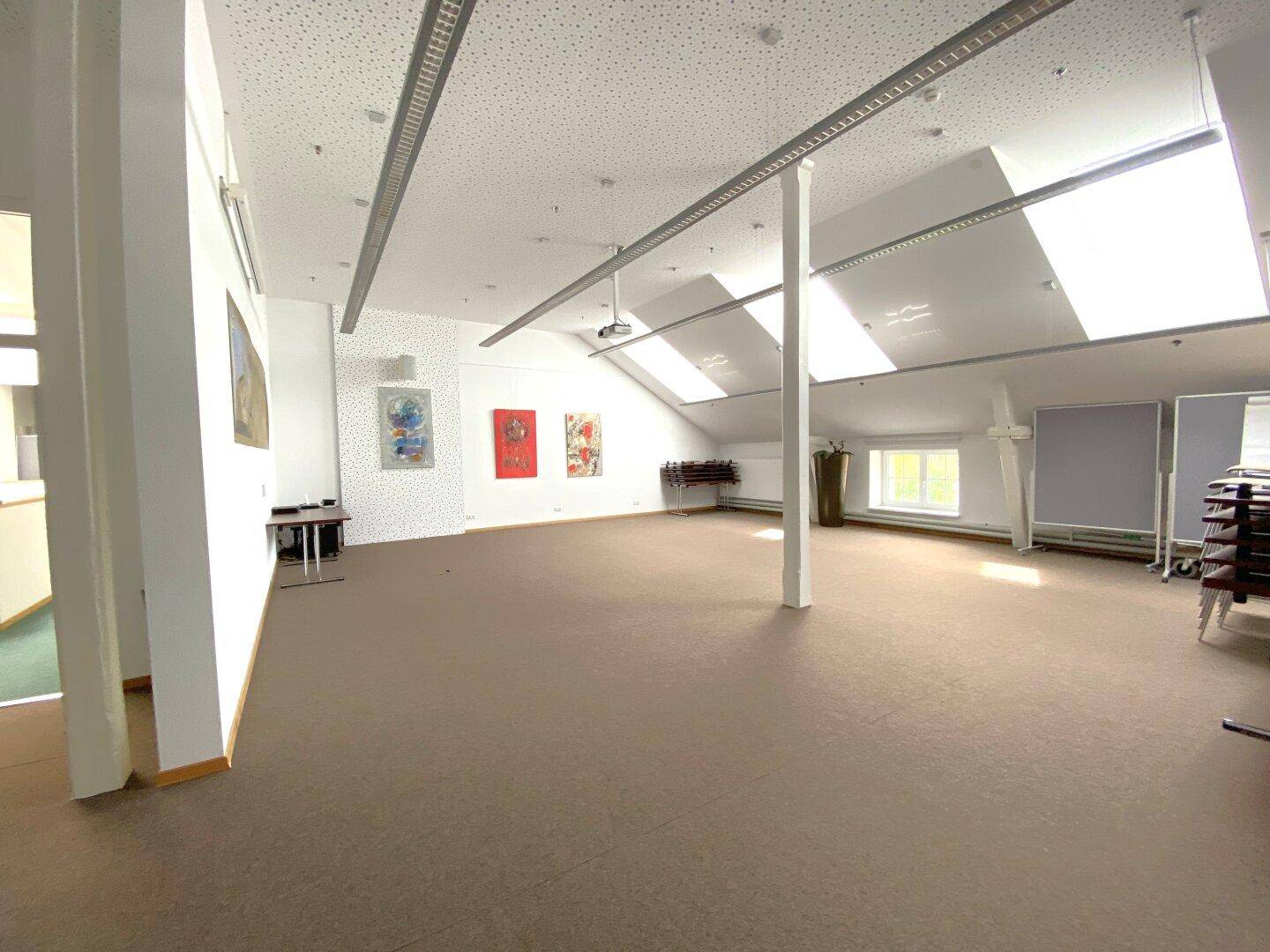 Großer Konferenzraum im Dachgeschoss mit Teppichboden, Dachfenstern, Projektor und Kunstwerken an den Wänden.
