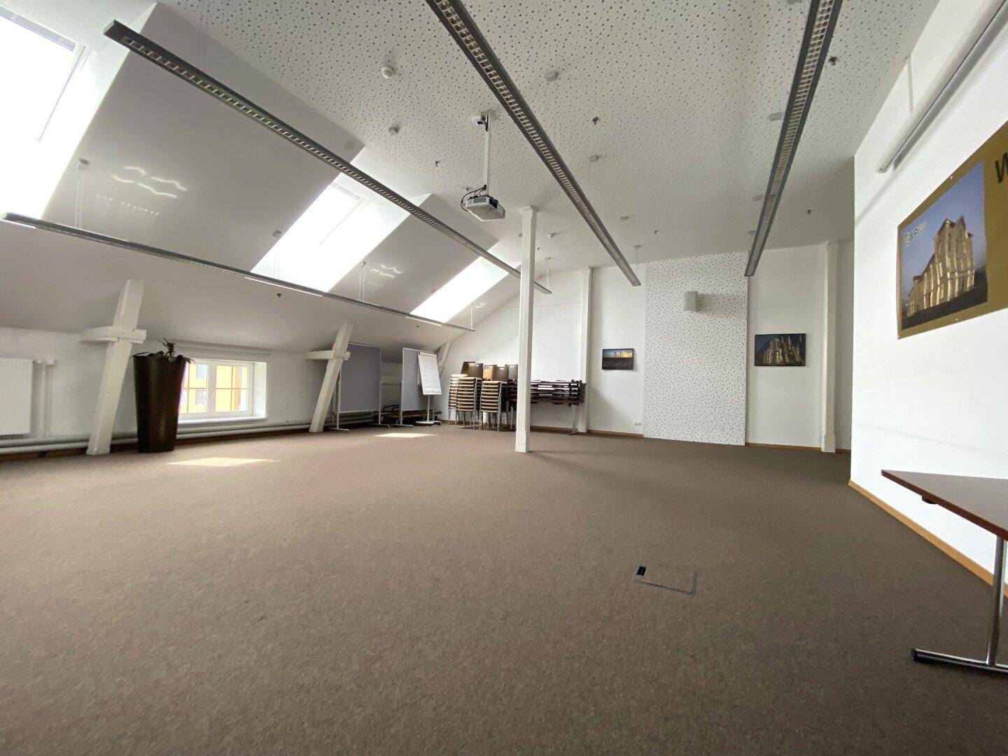 Großer Konferenzraum im Dachgeschoss mit Teppichboden, Dachfenstern, Projektor und Kunstwerken an den Wänden.