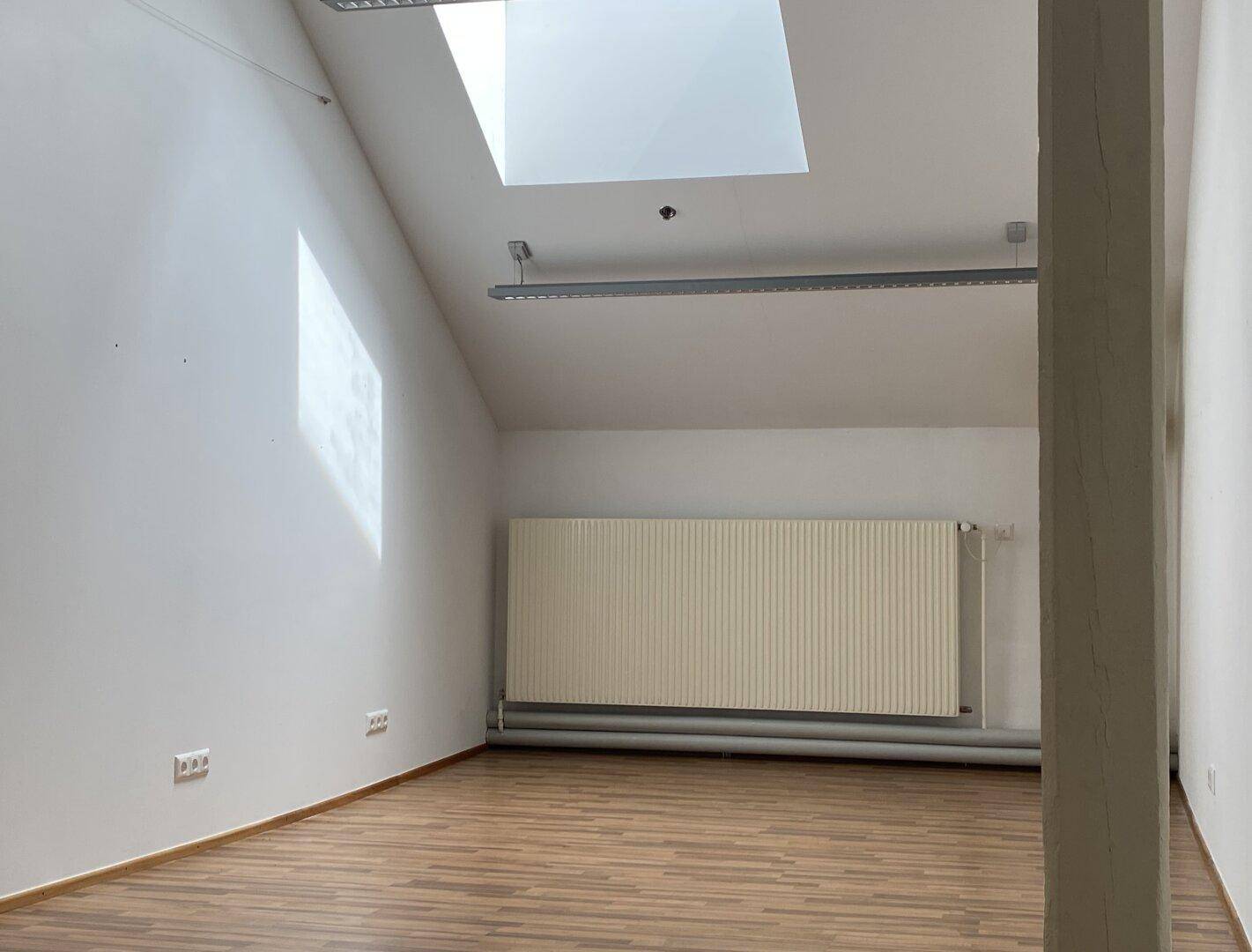Leerer Büroraum im Dachgeschoss mit Holzboden, Dachfenster und Heizkörper, bietet viel Tageslicht.
