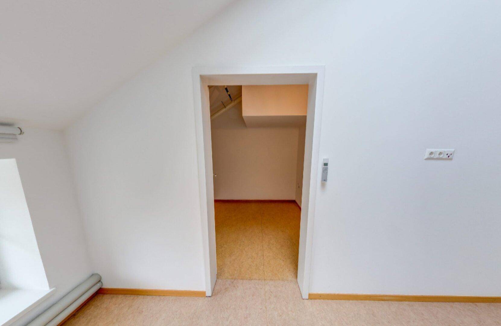 Türöffnung führt zu einem weiteren Raum mit Teppichboden und schrägen Wänden im Dachgeschoss.
