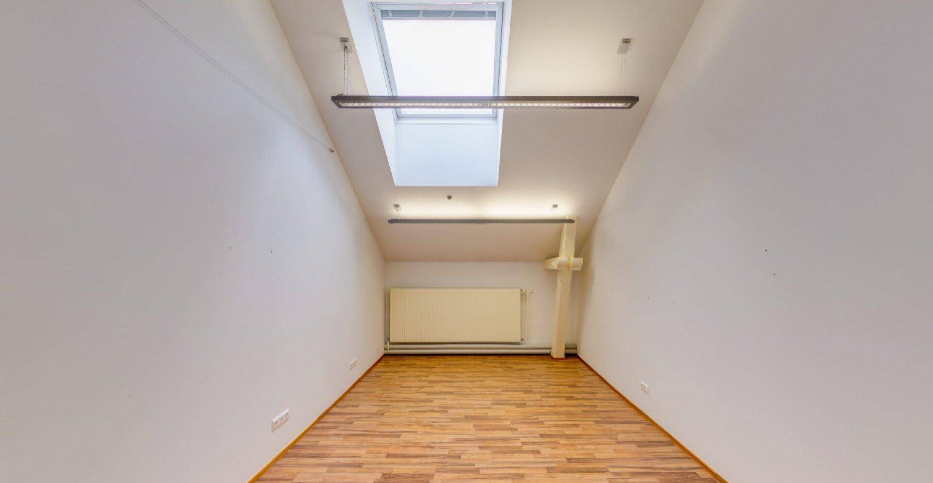 Heller Büroraum im Dachgeschoss mit Holzboden, Dachfenster und Heizkörper, bietet viel Tageslicht.