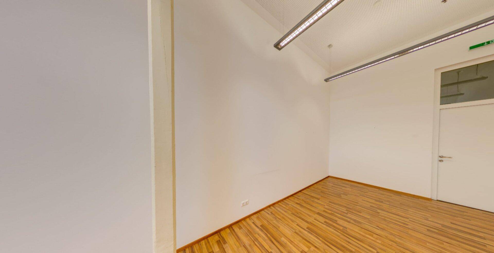 Leerer Büroraum mit hellem Holzboden und modernen Deckenleuchten, bereit zur individuellen Gestaltung.