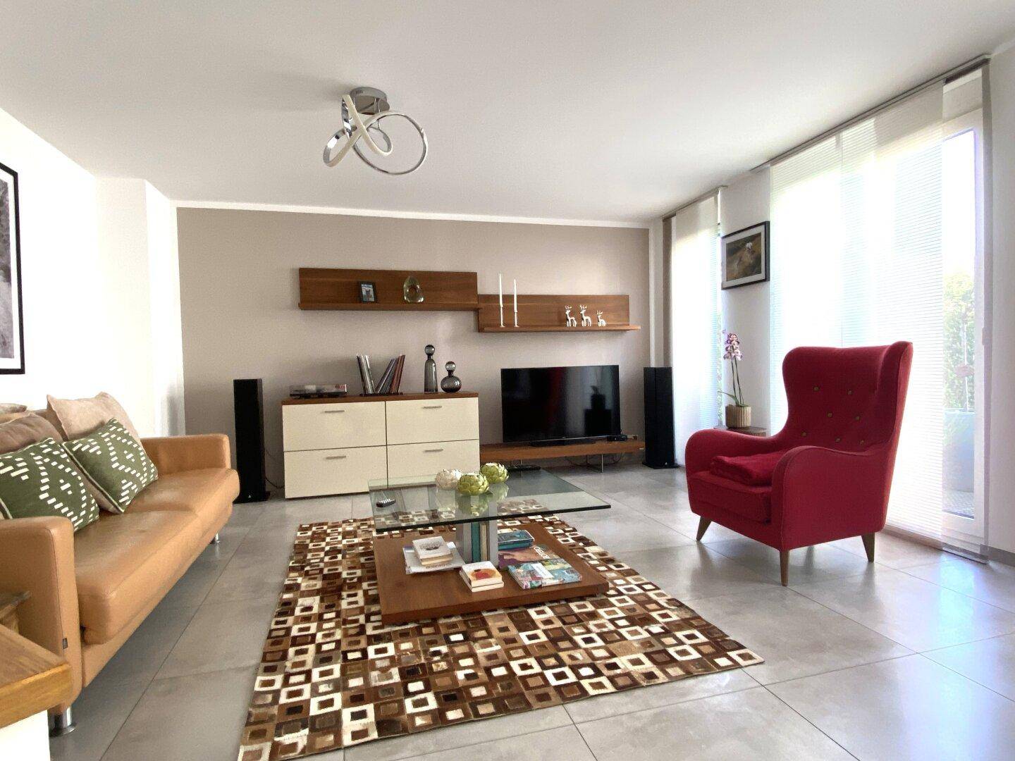 Helles Wohnzimmer mit beigem Sofa, rotem Sessel und großen Fenstern.