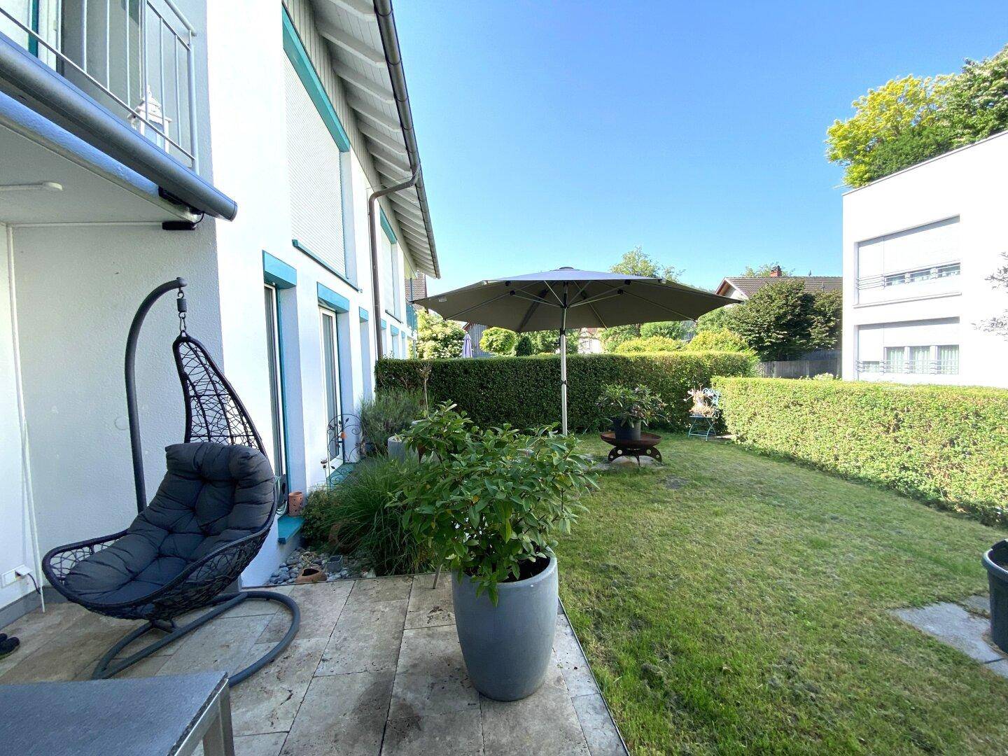 Sonnige Rasenfläche mit großem Sonnenschirm und Zugang zur Terrasse, umgeben von einer Hecke.