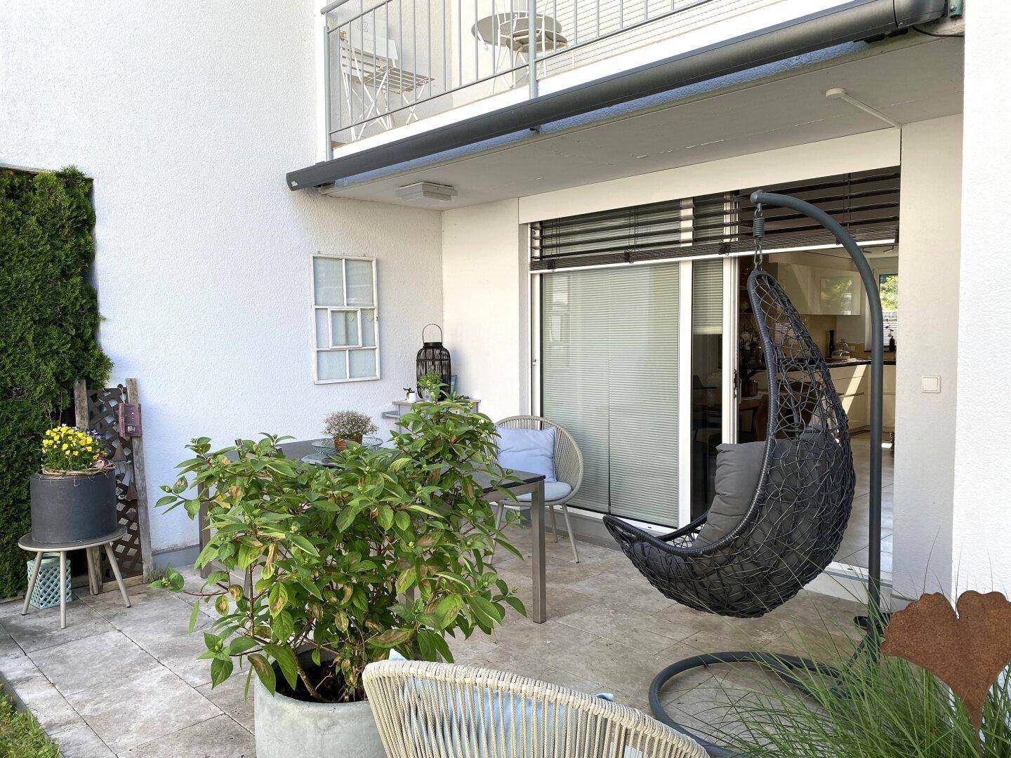 Gemütliche Terrasse mit Hängesessel und Esstisch, umgeben von Pflanzen und mit Zugang zum Innenbereich.