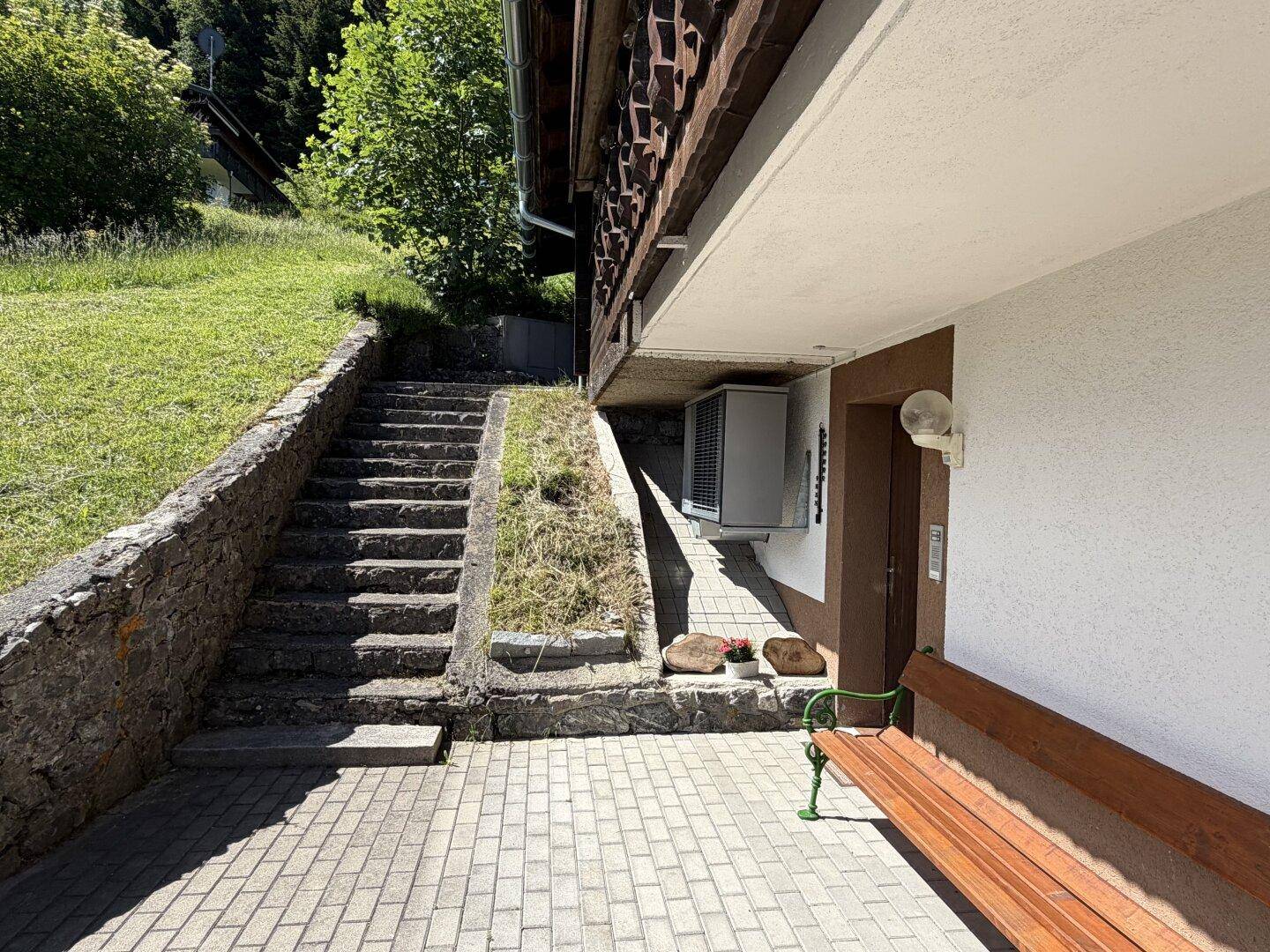 Eingangsbereich im Untergeschoss mit gepflastertem Vorplatz, Holzbank und Außeneinheit.