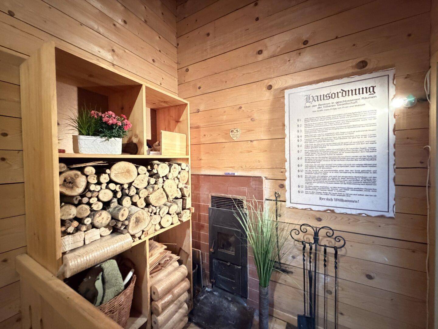 Gemütliche Ecke mit Holzofen, gestapeltem Brennholz und einer Hausordnung an der Wand.