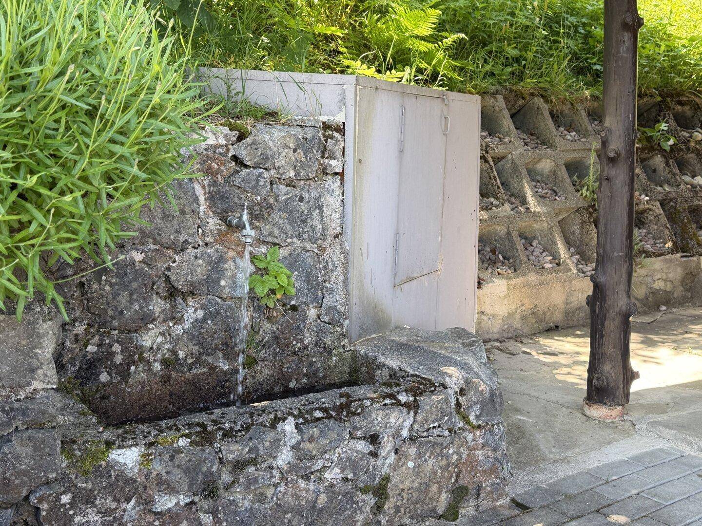 Rustikaler Steinbrunnen mit fließendem Wasser neben einer Natursteinmauer.