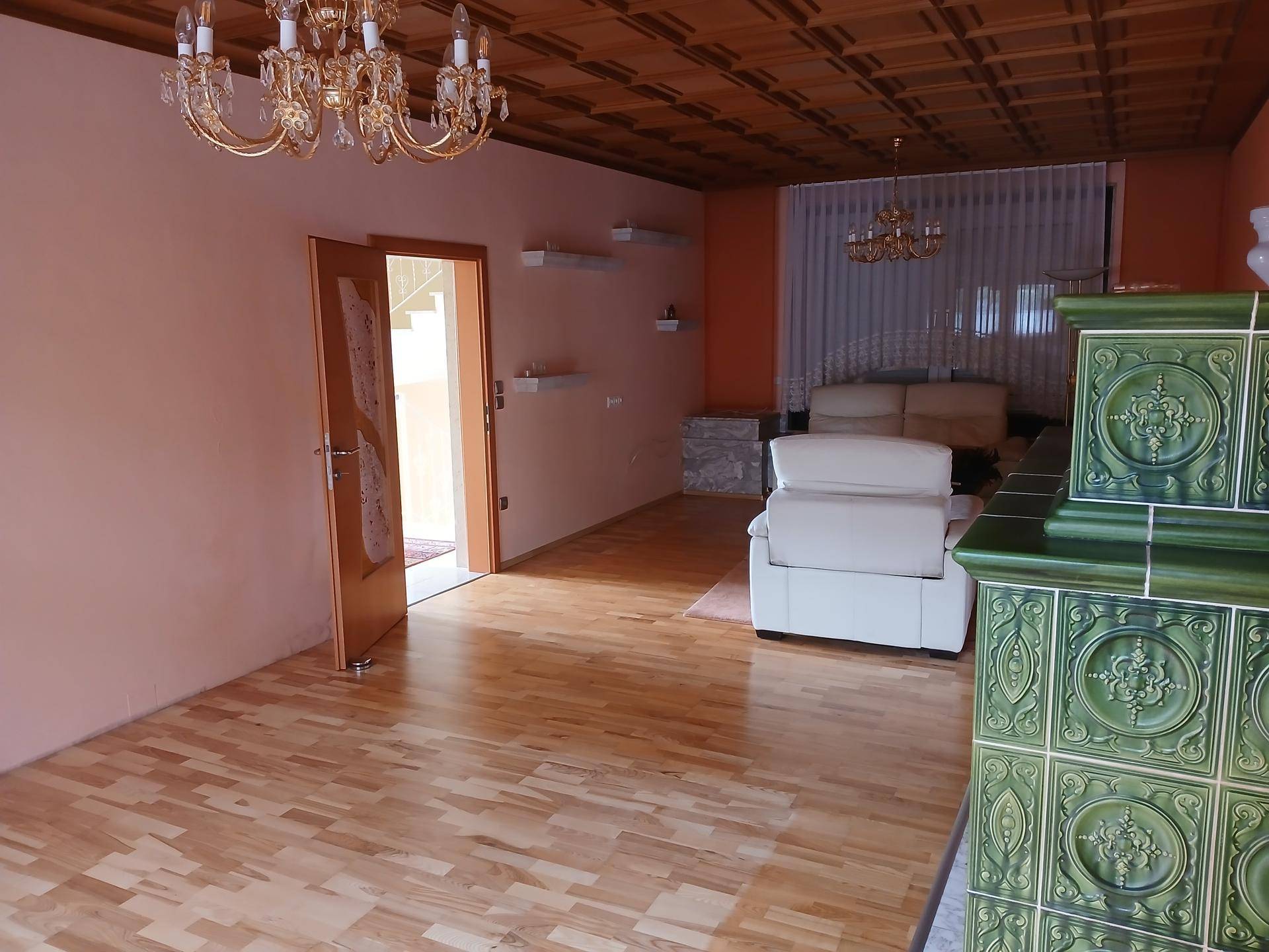 Geräumiges Wohnzimmer mit Kachelofen, Holzkassettendecke und Zugang zu weiteren Räumen.