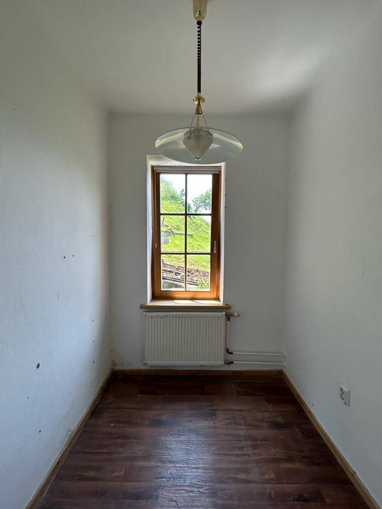 Kleiner, schmaler Raum mit Laminatboden, Fenster und Heizkörper, beleuchtet von einer Pendelleuchte.