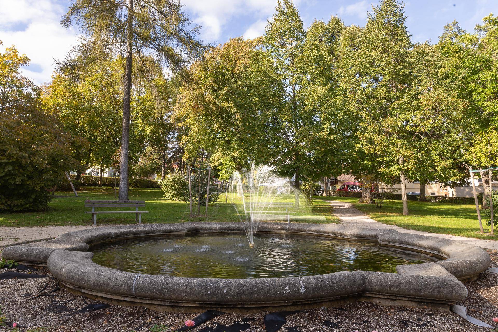 Ein idyllischer Park mit einem Steinbrunnen und vielen Bäumen an einem sonnigen Tag.