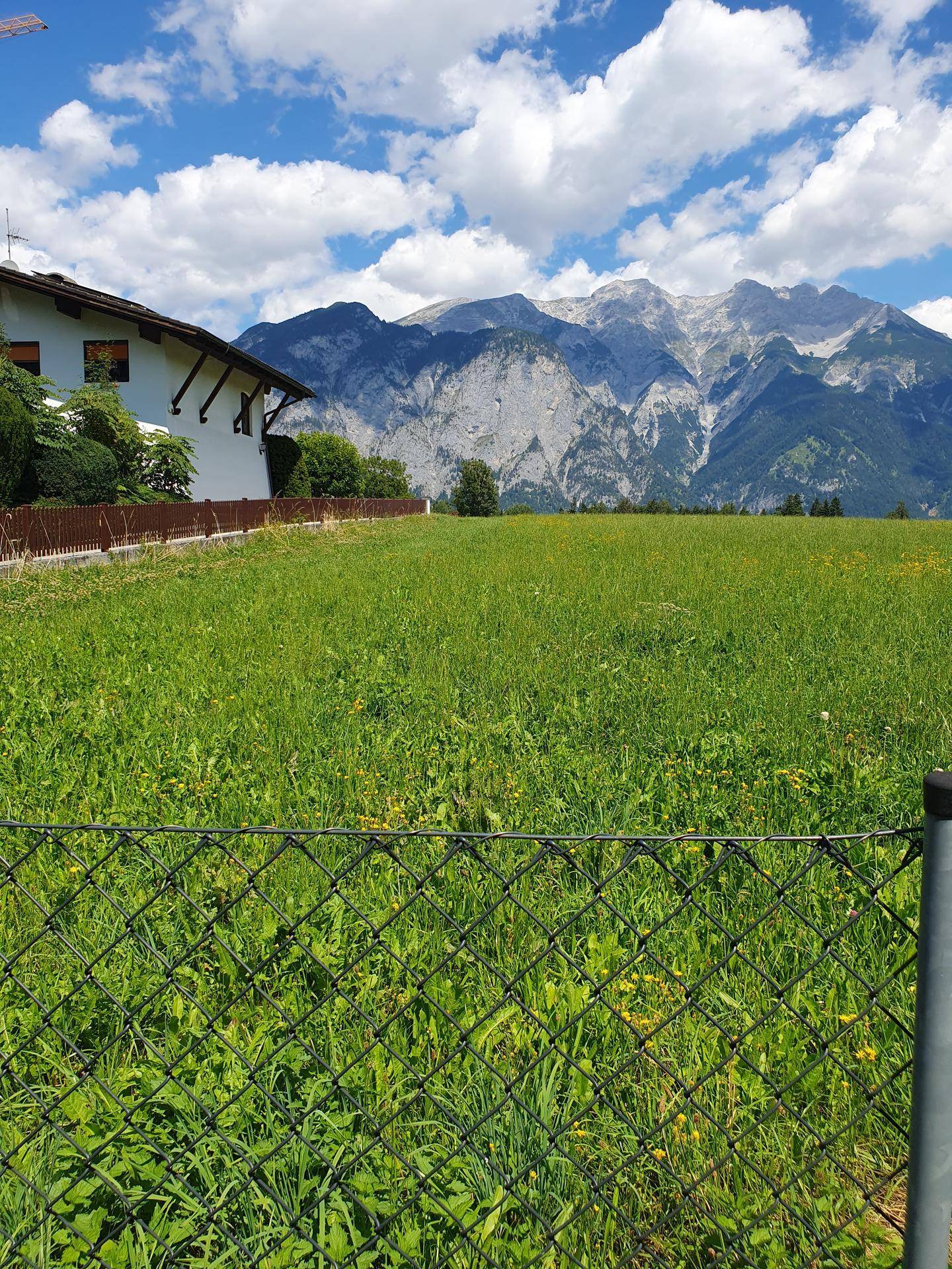 Grünes Feld mit einem Haus am Rand und einer beeindruckenden Bergkulisse unter strahlend blauem Himmel.