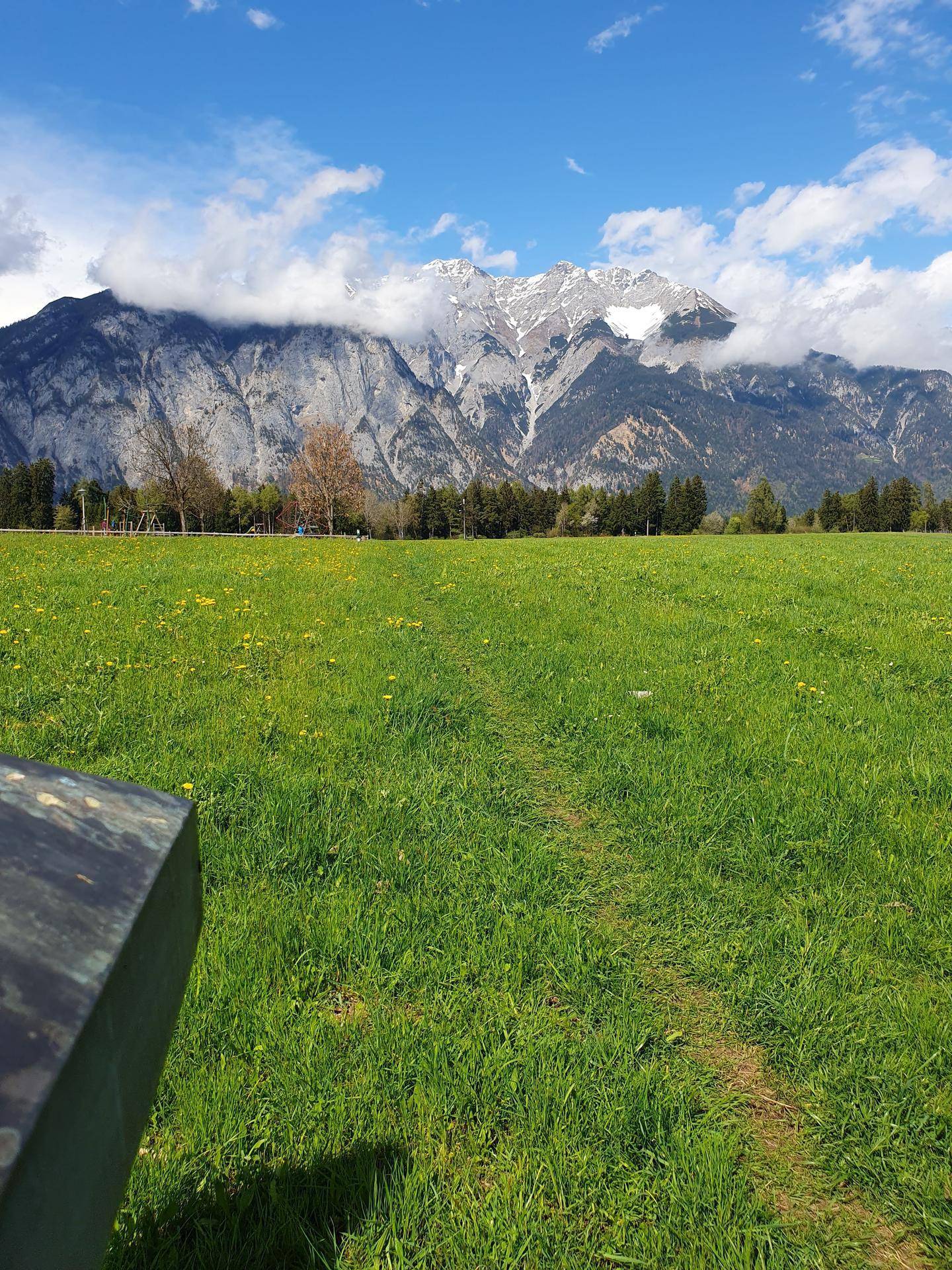 Sonnenbeschienenes grünes Feld mit majestätischen Bergen, deren Gipfel von Wolken umhüllt sind, unter blauem Himmel.