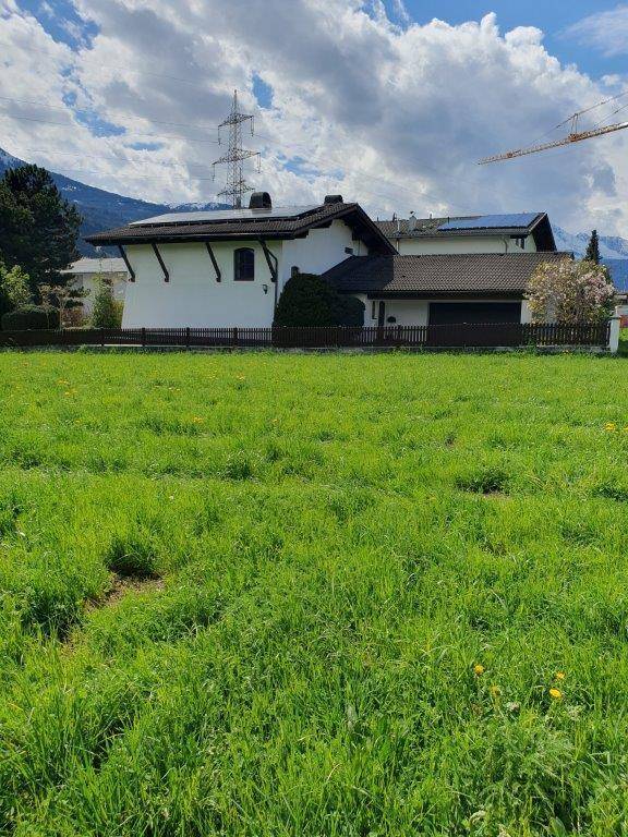 Weitläufiges grünes Feld vor einem traditionellen Haus mit Solarpaneelen auf dem Dach unter bewölktem Himmel.