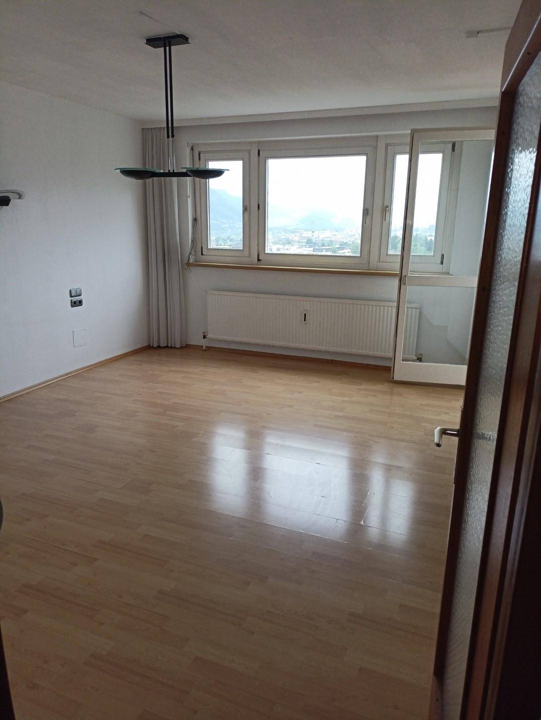 Helles, leeres Wohnzimmer mit Laminatboden, großen Fenstern und Blick auf die Stadt.