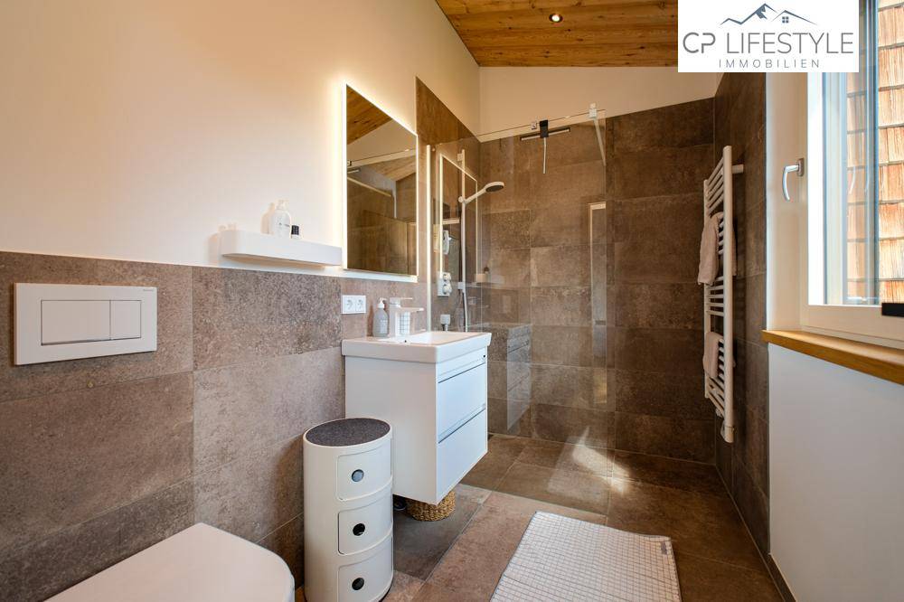 Elegantes Badezimmer mit braunen Fliesen, großer Dusche und beleuchtetem Spiegelschrank.