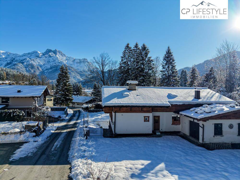 Traditionelles Haus mit schneebedecktem Dach und Vorgarten, umgeben von Bergen und Bäumen.