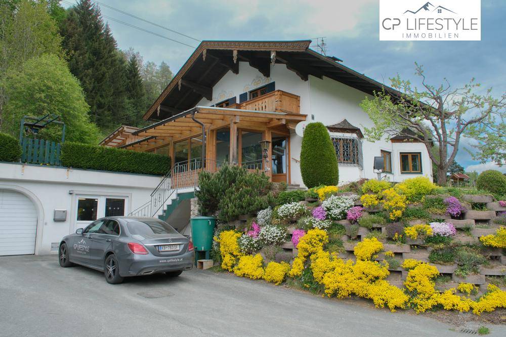 Alpines Haus mit Garage, Auffahrt und blühenden Blumenbeeten in bergiger Landschaft.