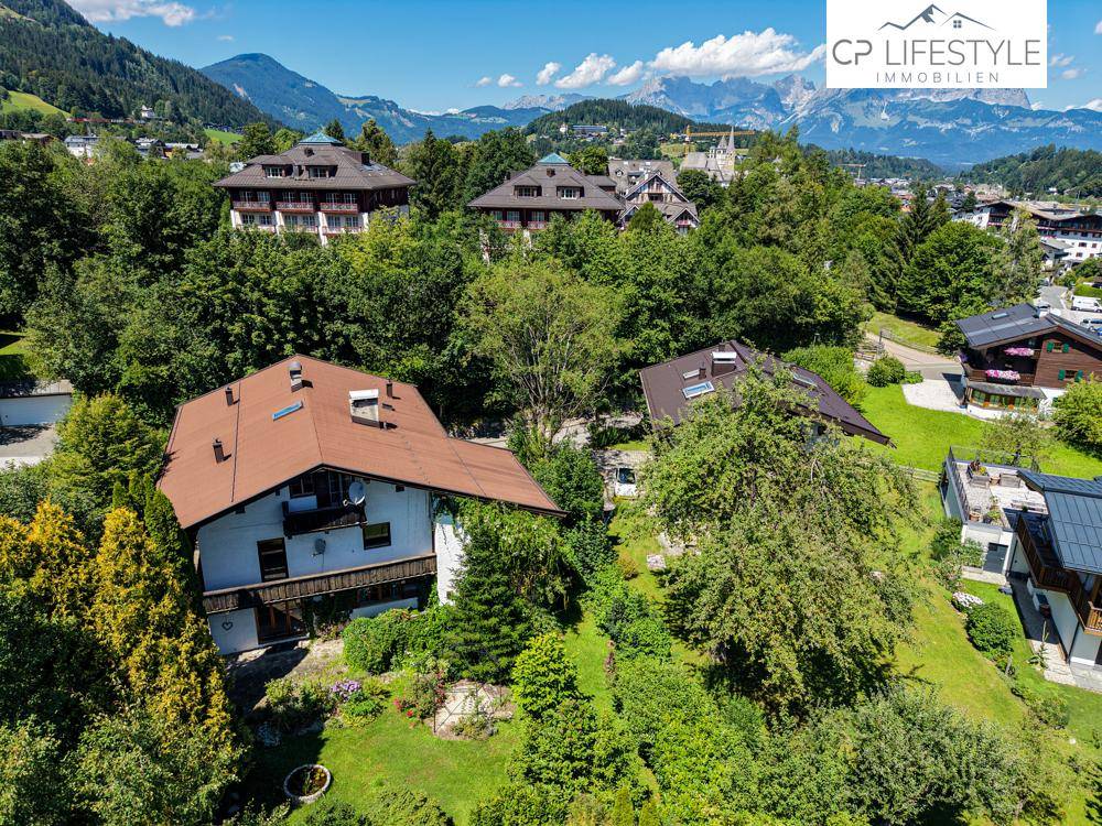 Luftaufnahme eines Wohngebiets mit Häusern, umgeben von üppiger Vegetation und Bergen im Hintergrund.