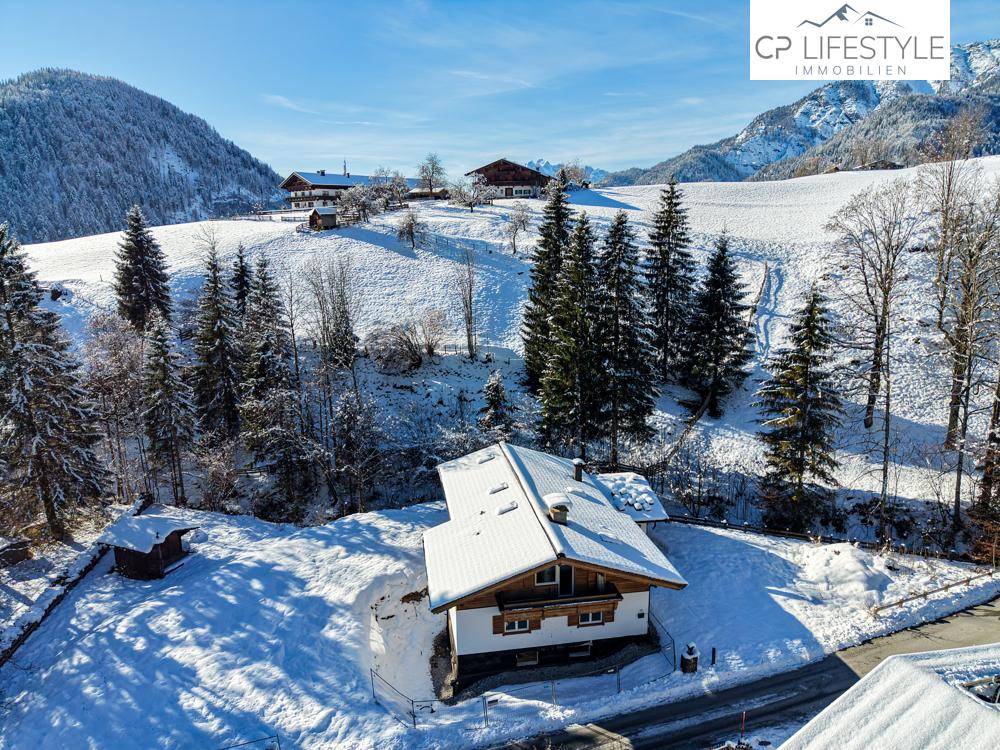 Einzelnes Haus in einer verschneiten Berglandschaft, umgeben von Nadelbäumen und einem klaren Himmel.