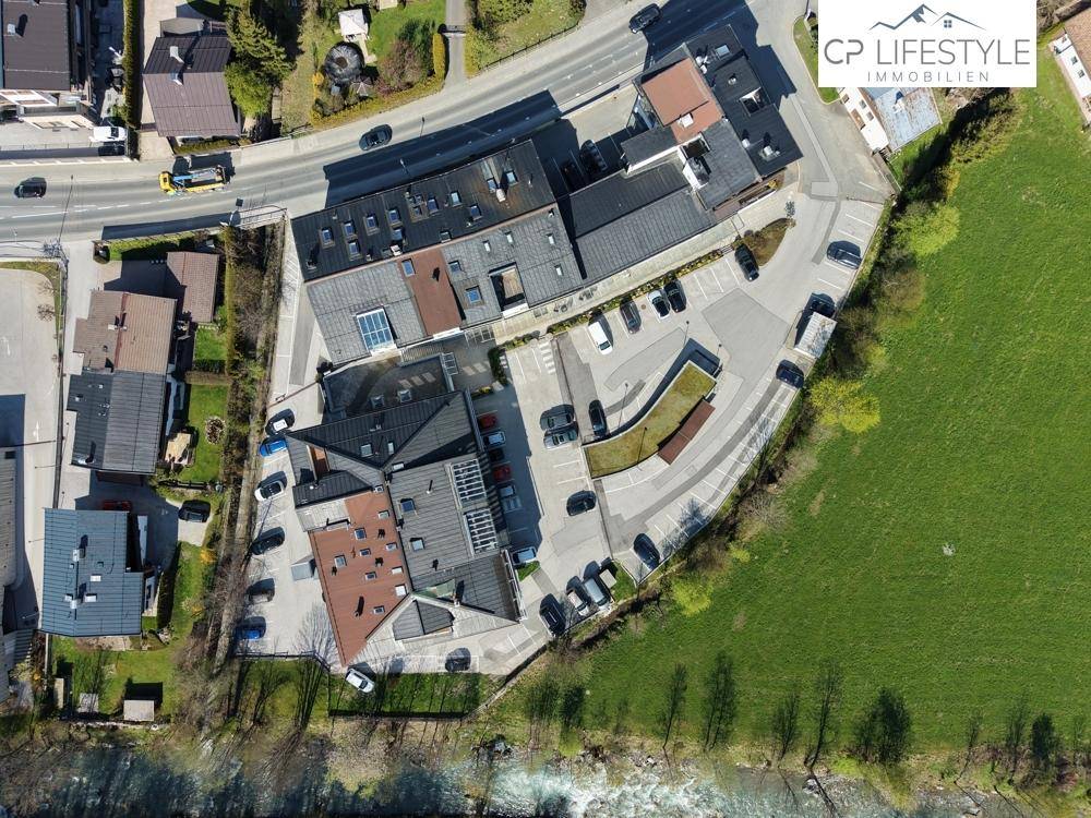 Top-Down-Luftaufnahme eines Gebäudekomplexes mit Parkplätzen, umgeben von Grünflächen und einem Fluss.