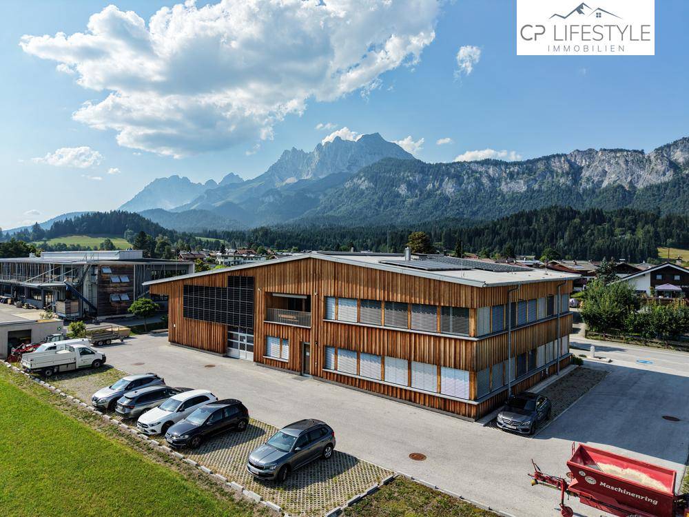 Luftaufnahme eines modernen Geschäftsgebäudes mit Holzfassade, Parkplätzen und einer beeindruckenden Bergkulisse.
