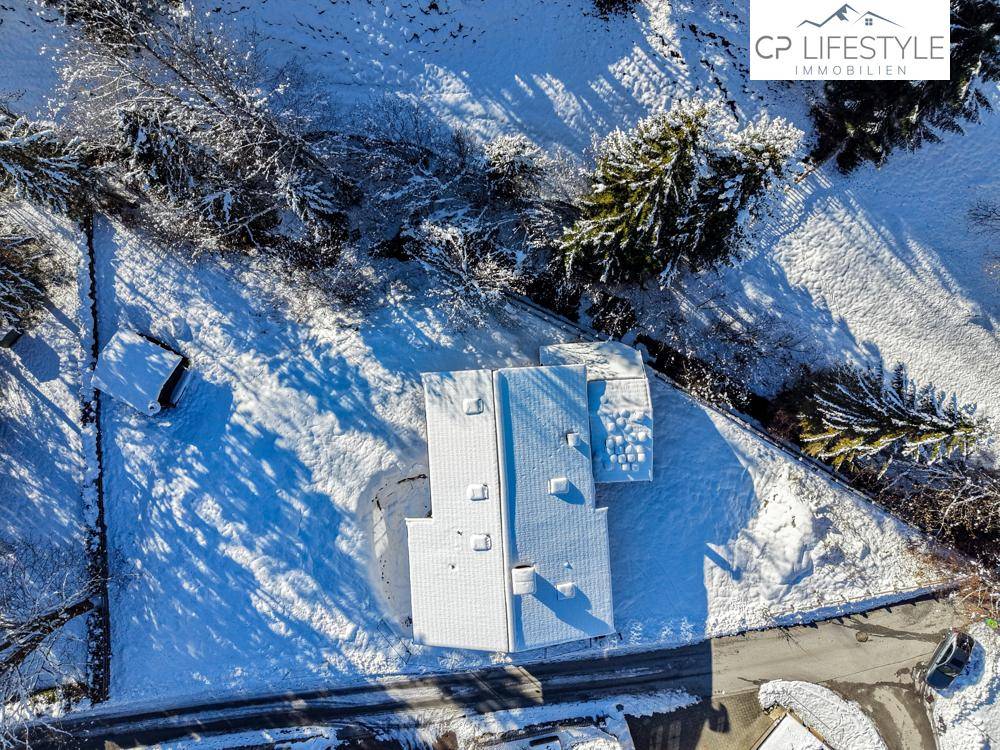 Draufsicht auf ein schneebedecktes Haus mit umliegenden Bäumen und Wegen in winterlicher Umgebung.