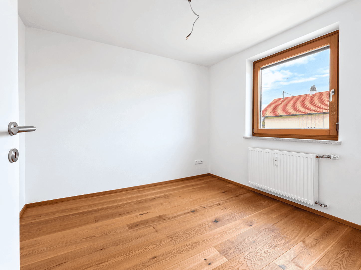 Leeres Schlafzimmer mit Holzboden und Fenster mit Blick auf Nachbardächer.