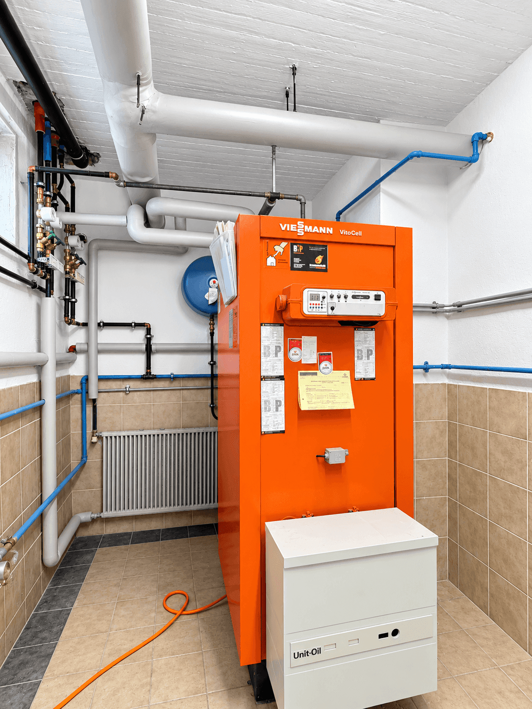 Heizungsraum mit großem orangefarbenem Ölkessel und Rohrleitungen.