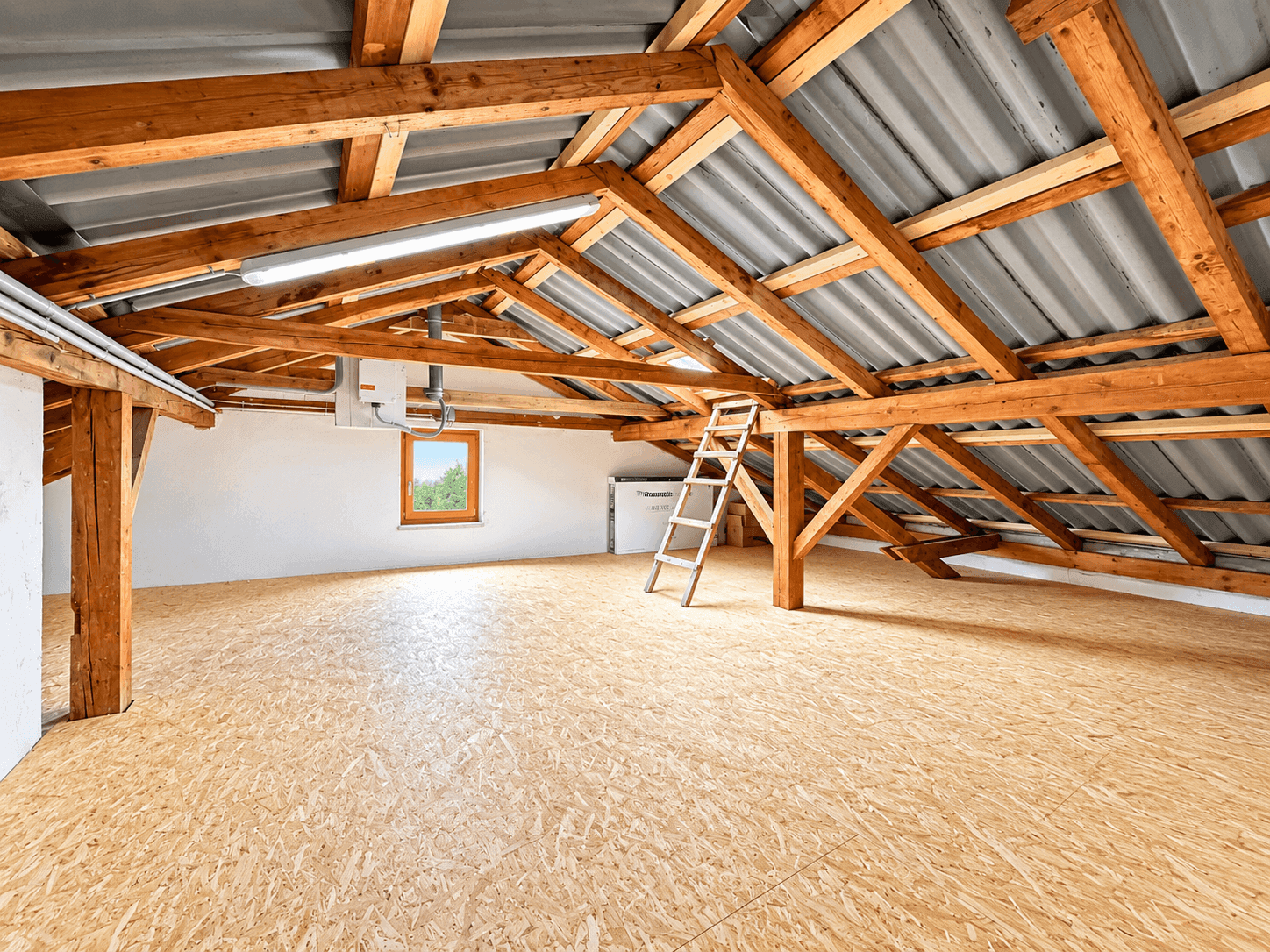 Geräumiger Dachboden mit sichtbaren Holzbalken, Fenster und Holzspanplattenboden.