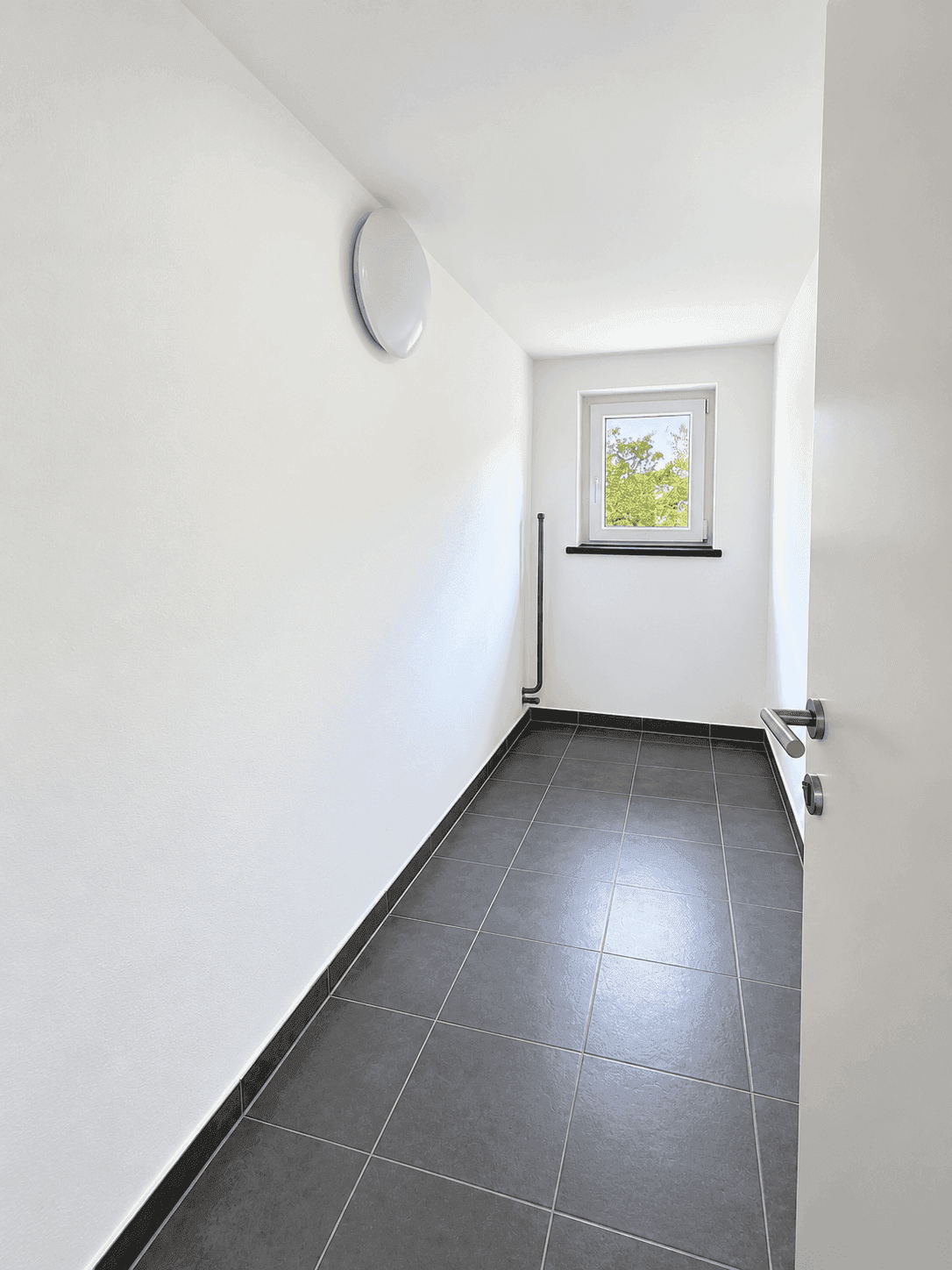 Schmaler Flur mit Fliesenboden und Fenster, ideal als Abstellbereich.