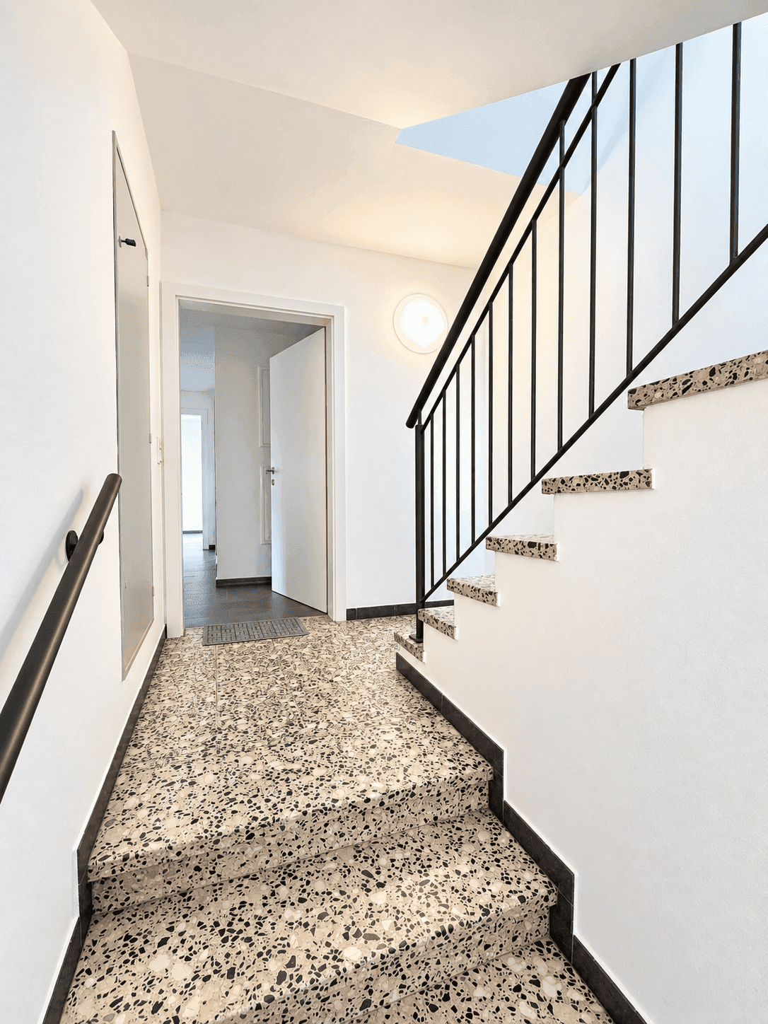 Heller Eingangsbereich mit Terrazzoboden und Treppe mit schwarzem Geländer.